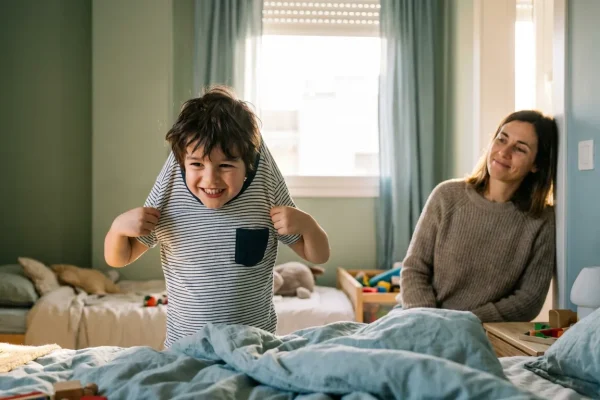 Niño poniéndose la camiseta él solo mientras su madre observa con paciencia desde la puerta